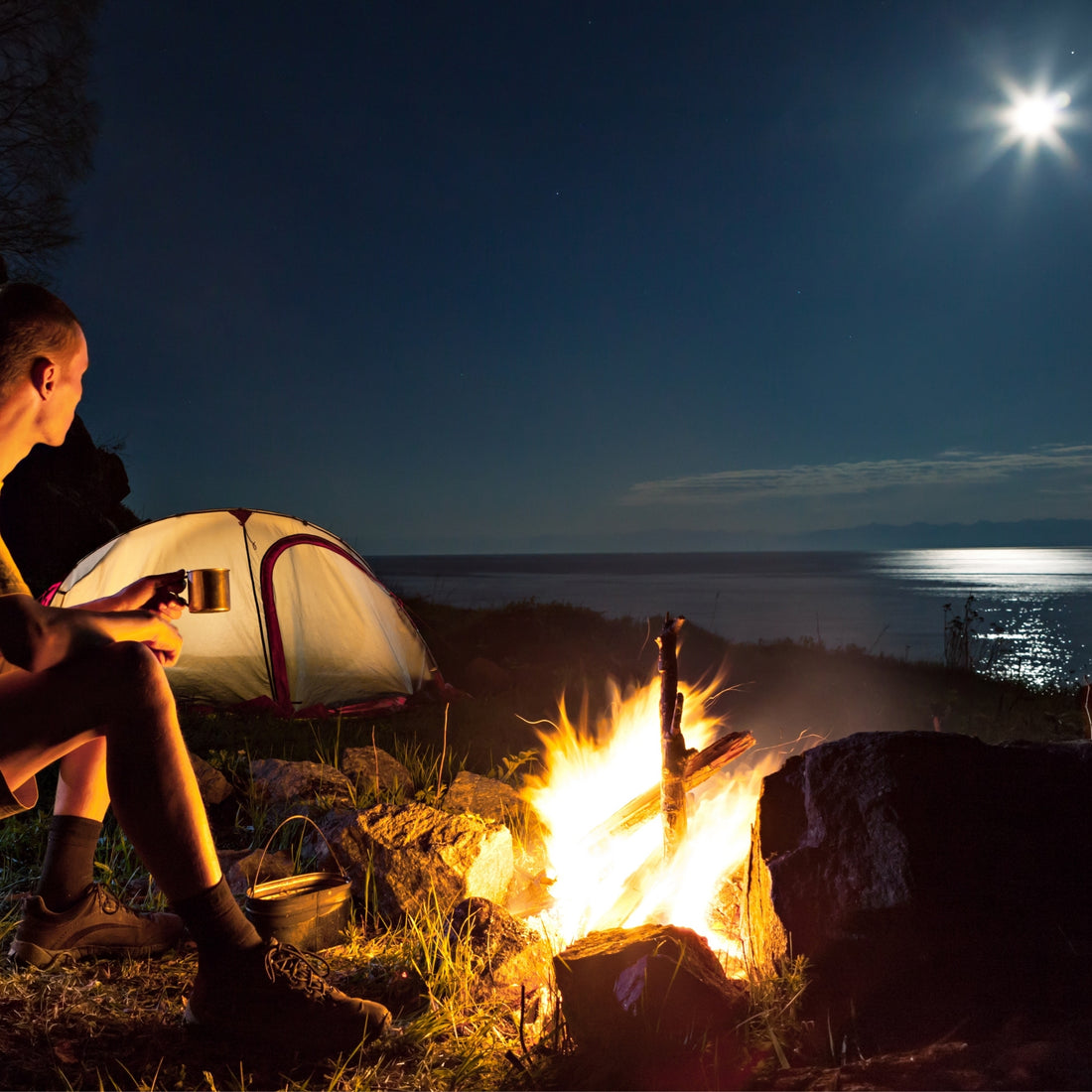 Man sitting next to camp fire near tent overlooking water